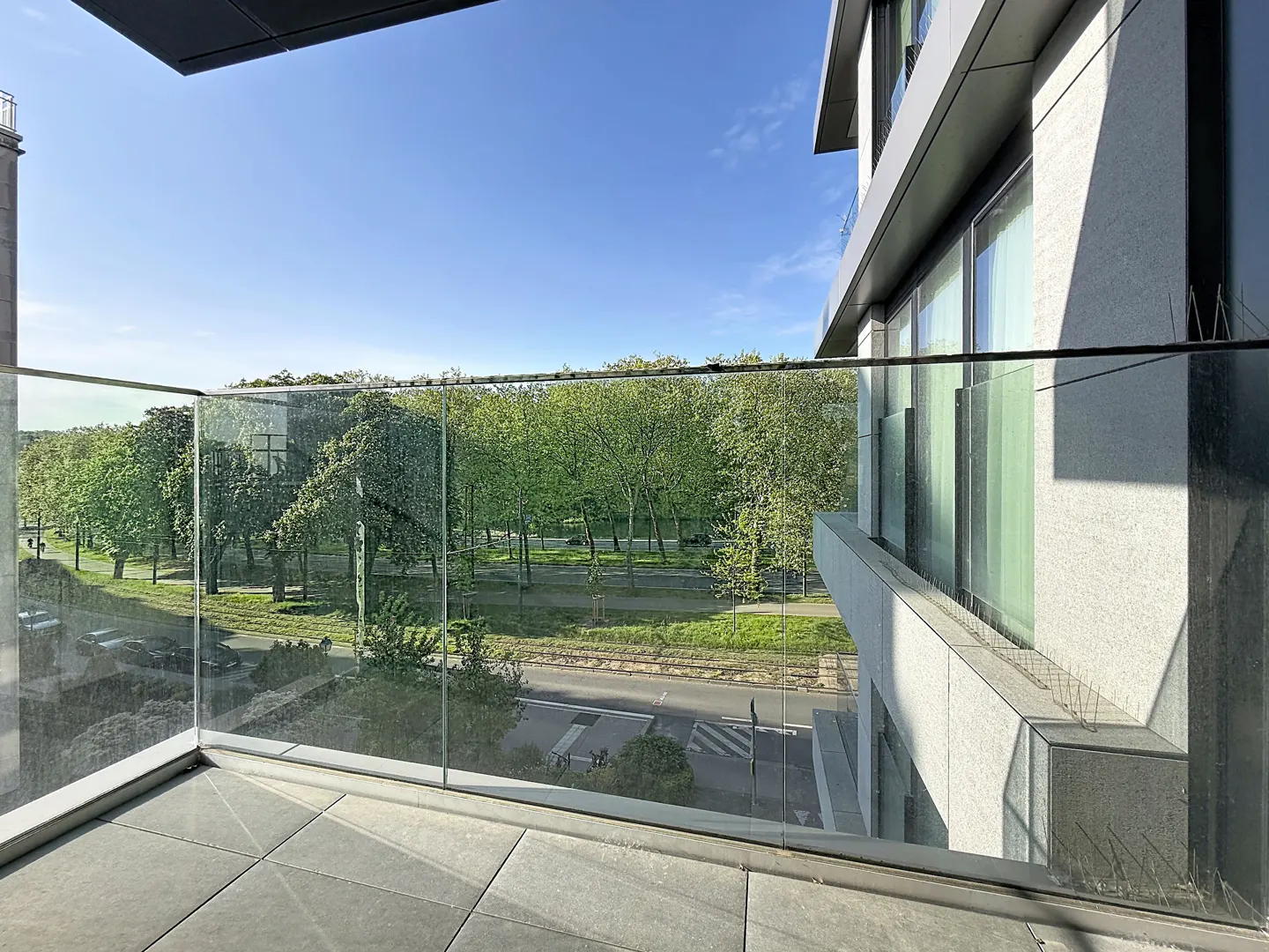 Balcony view of a tree-lined street. Glass railing, gray stone tiles, and a modern building are visible. Blue sky.