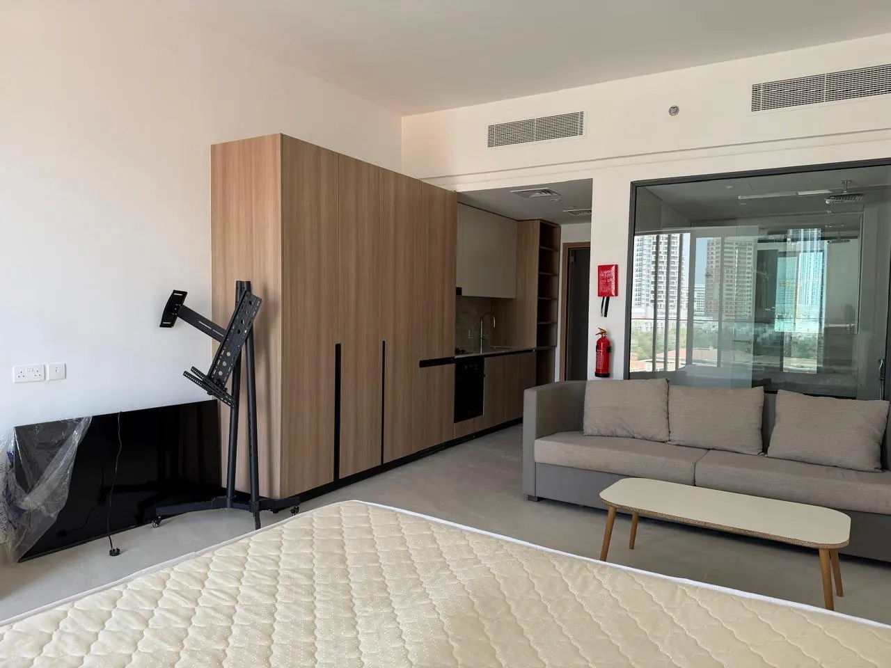 Studio apartment with a mattress in the foreground, a gray sofa, and a kitchen area with wood cabinets in the background.