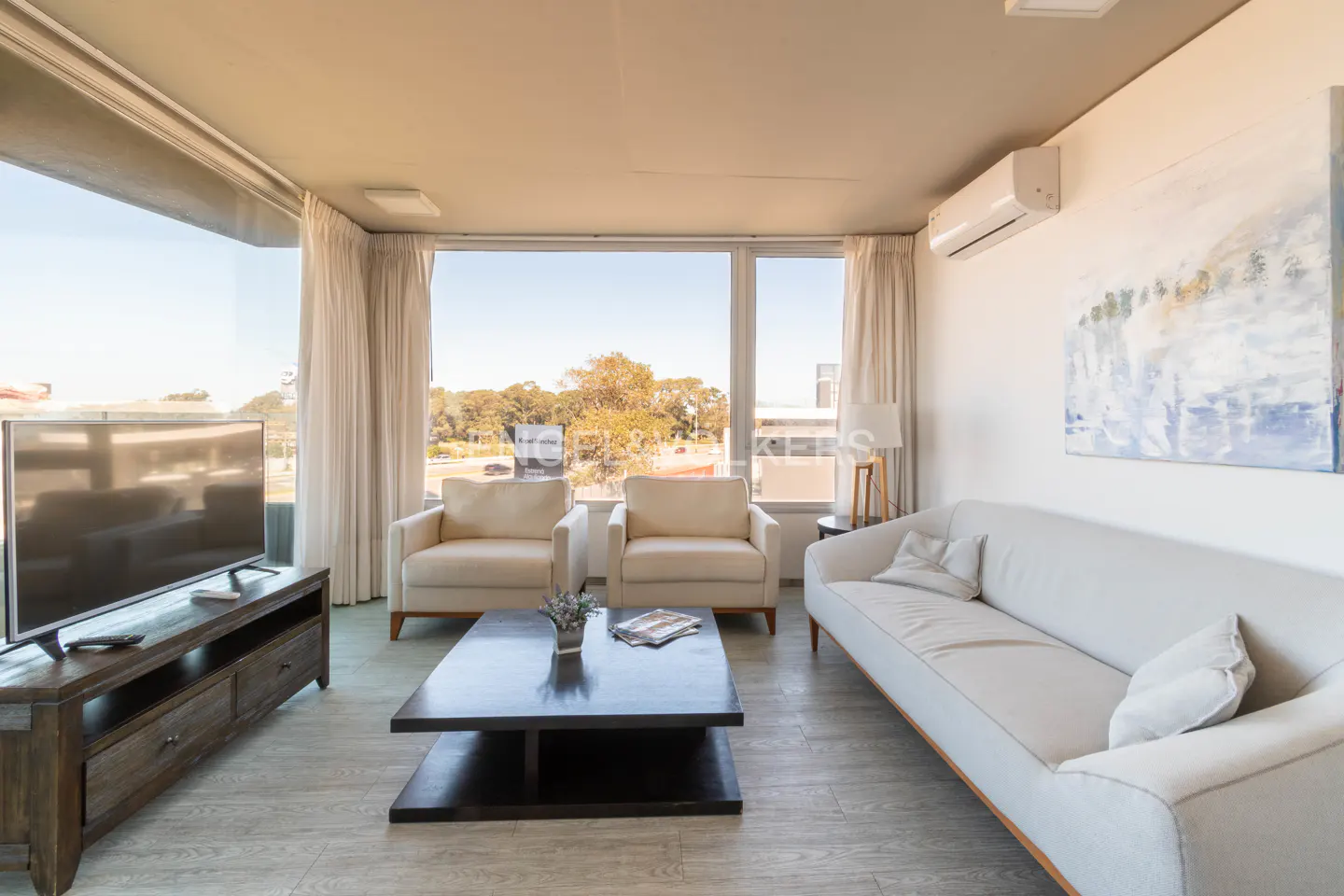 Bright living room with a white sofa, two armchairs, a dark coffee table, and a TV with a view of trees outside.
