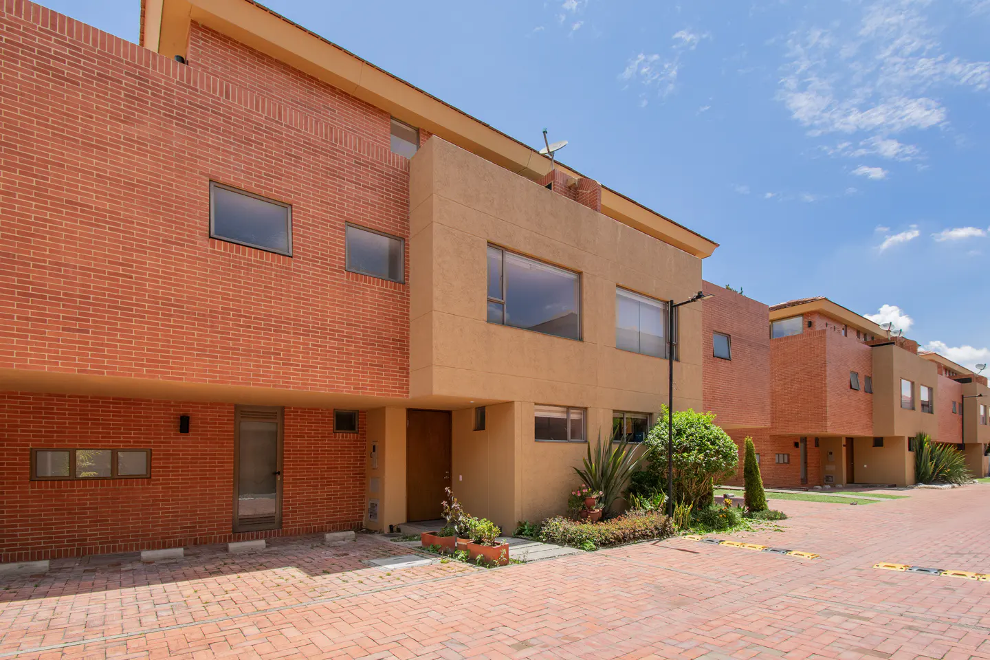 Row of modern townhouses with brick and tan facades under a blue sky. Brick paved driveway and small garden in front.