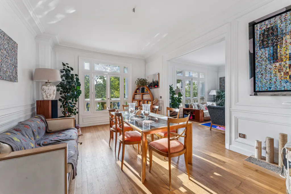 Bright dining room with wood floors, a glass-topped table with orange chairs, and a white sofa. Large windows offer a view of greenery.