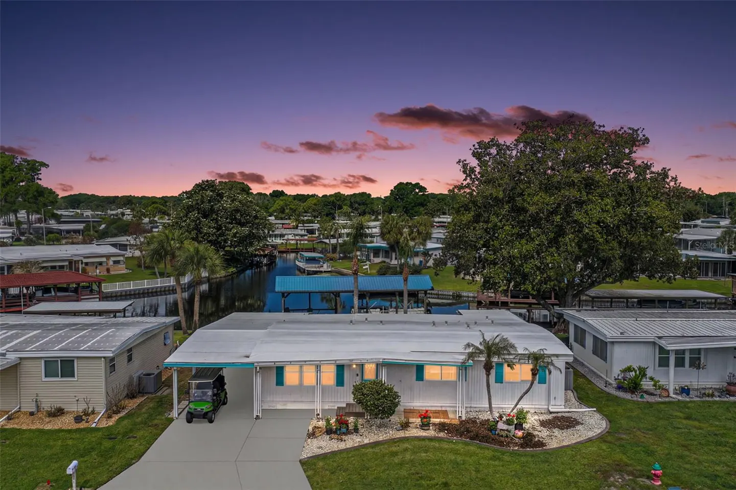 Aerial view of a white single-story home with turquoise shutters and a green golf cart in the driveway. A canal runs behind the house. Sunset sky.