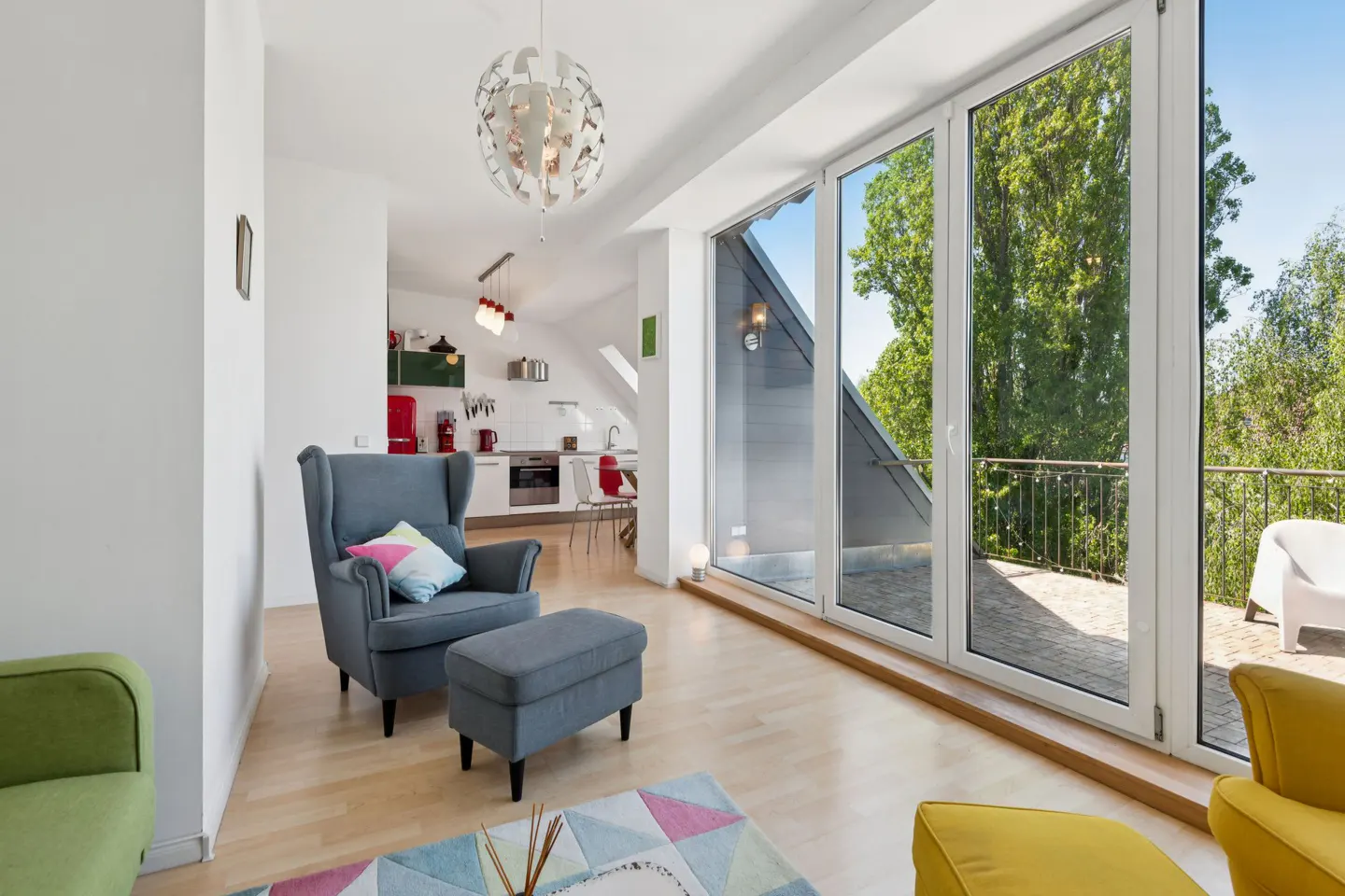 Bright, modern living room with a blue armchair, open kitchen, and large windows overlooking green trees.