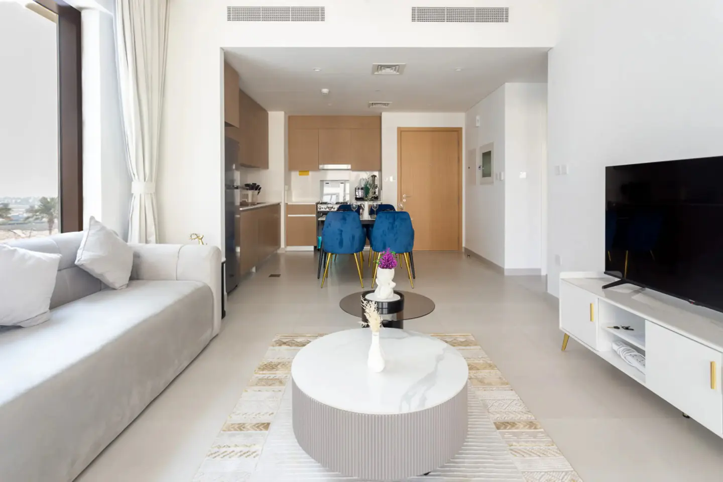 Bright, modern apartment interior with a gray sofa, round coffee tables, and blue dining chairs. A TV stands on a white console.