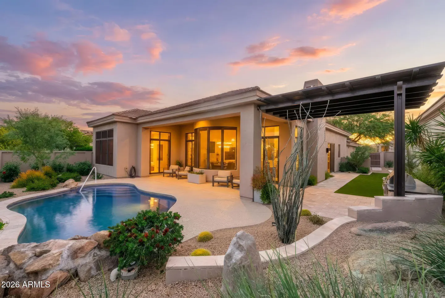 Beige house with a blue pool, patio furniture, and desert landscaping under a pink and purple sunset sky.