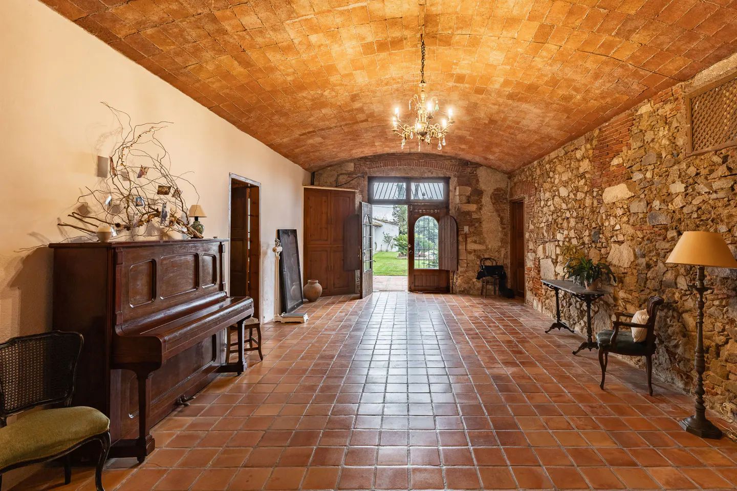 Interior view of a room with terracotta tile floor and ceiling, stone wall, piano, and open doors to a garden.