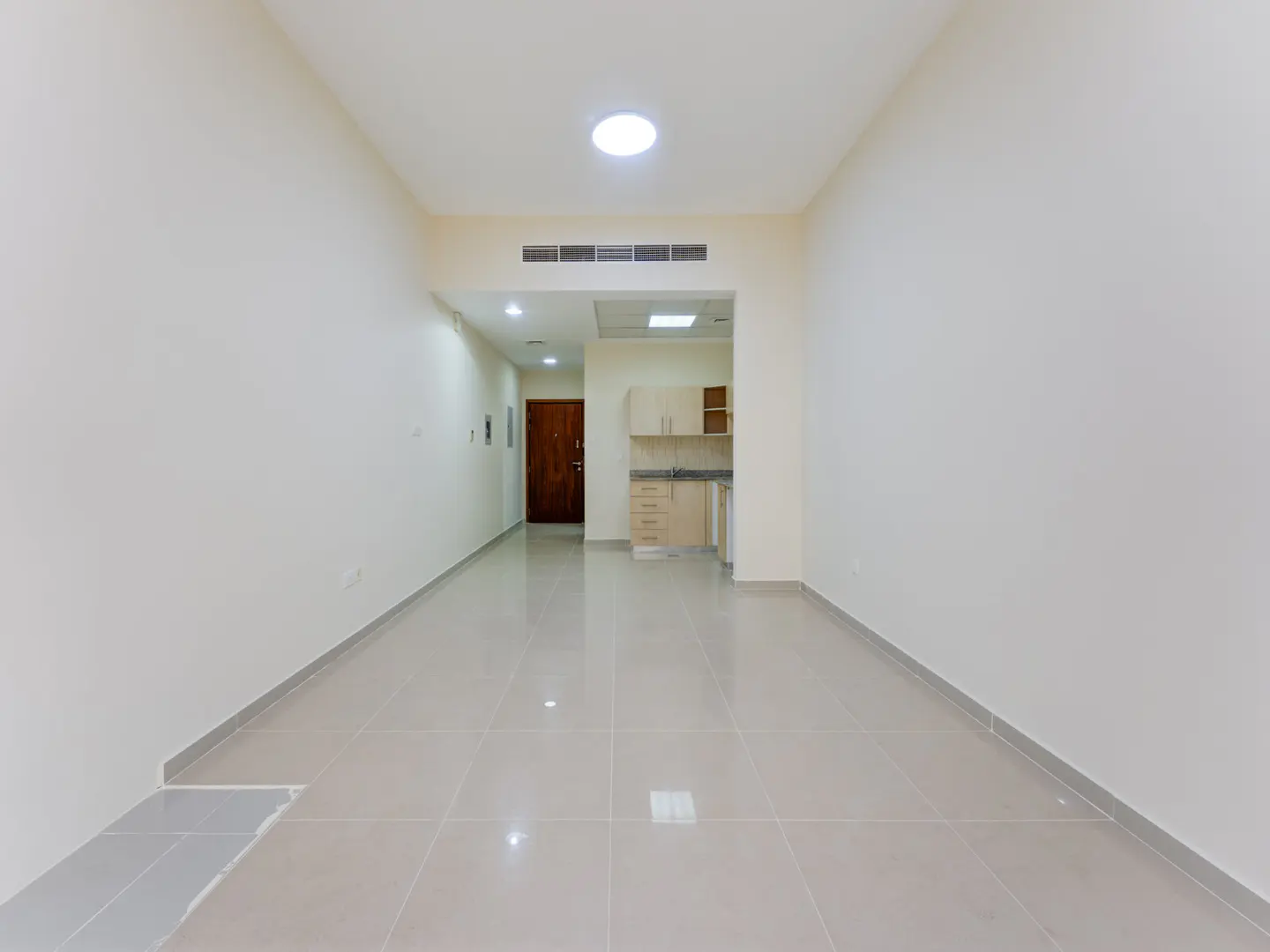 Empty, long room with white walls, light tile floors, a small kitchen area in the back, and a brown wooden door.