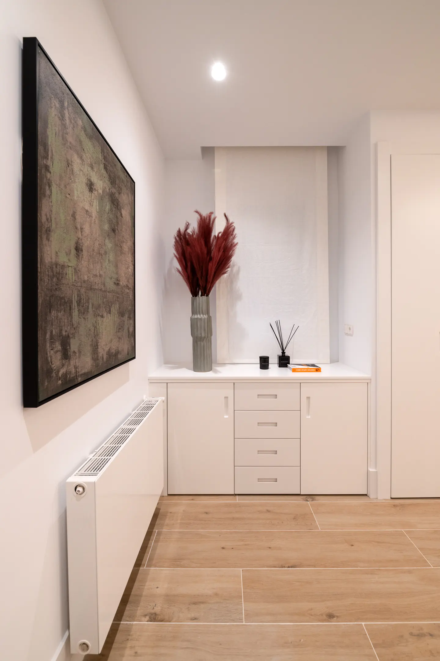 A modern room with wood floors, white walls, and a large abstract painting. A white cabinet holds a vase of red pampas grass.
