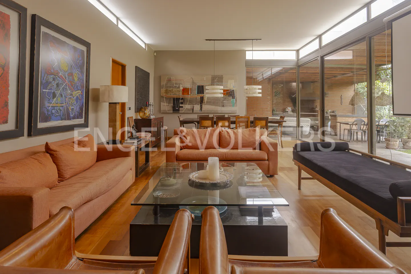 A modern living room with orange sofas, a glass coffee table, and large windows. Artwork adorns the walls.