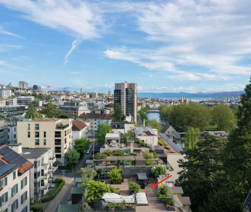 Cityscape view from a rooftop terrace with green plants, table and chairs, under a blue sky with clouds.