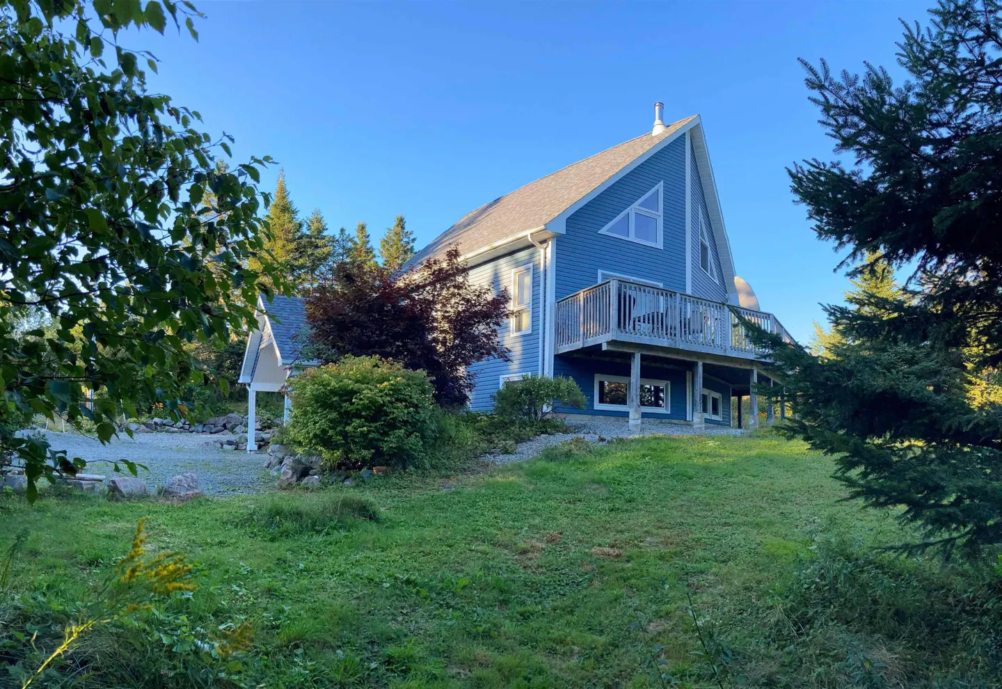 Blue A-frame house with a deck, surrounded by green grass and trees under a clear blue sky.