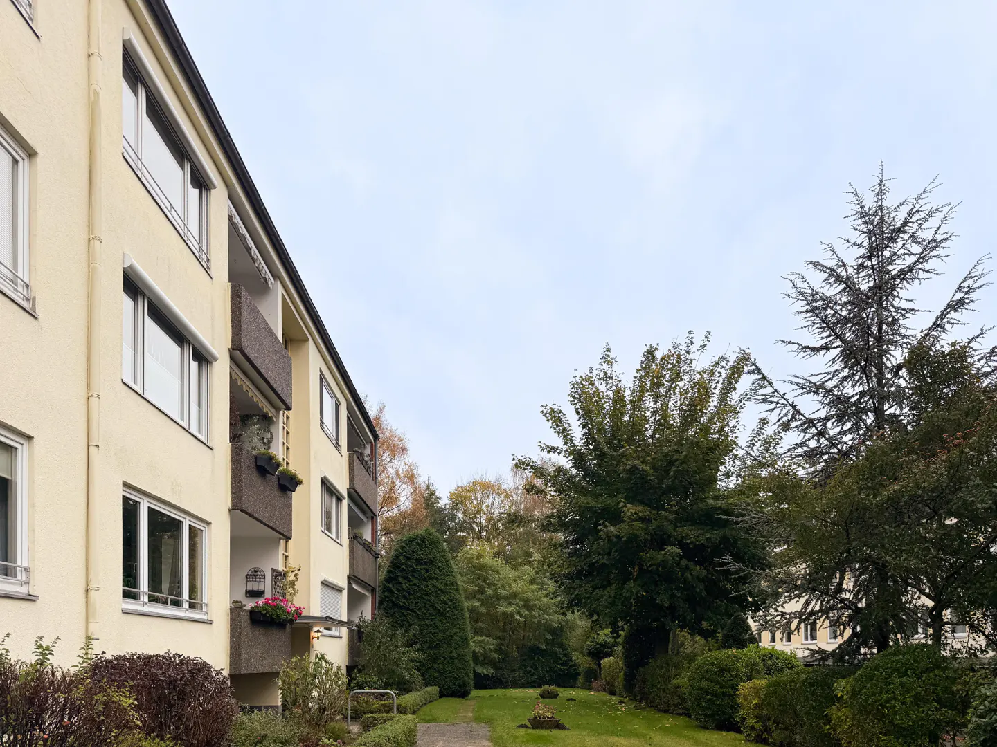 A low-rise apartment building with balconies overlooks a green lawn and trees. The building is light yellow with white-framed windows.