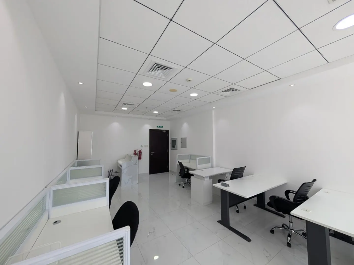 Bright, empty office space with white walls, tiled floor, and desks with black chairs.