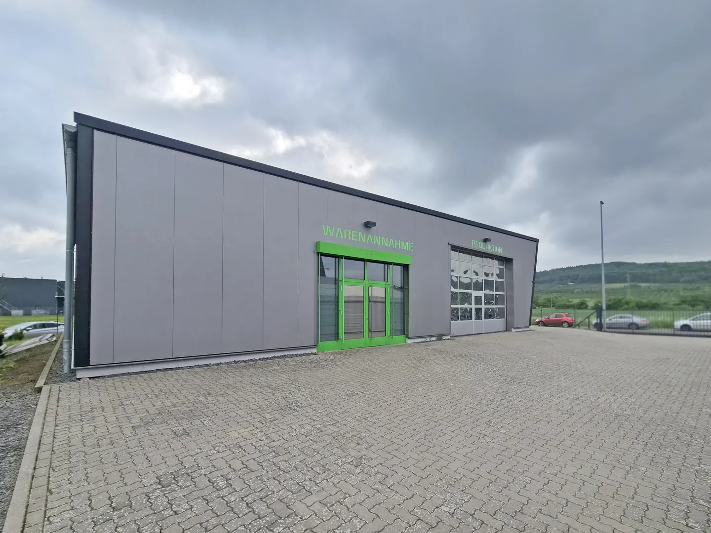 Exterior view of a gray warehouse with green doors and "Warenannahme" sign. Cars are parked outside.