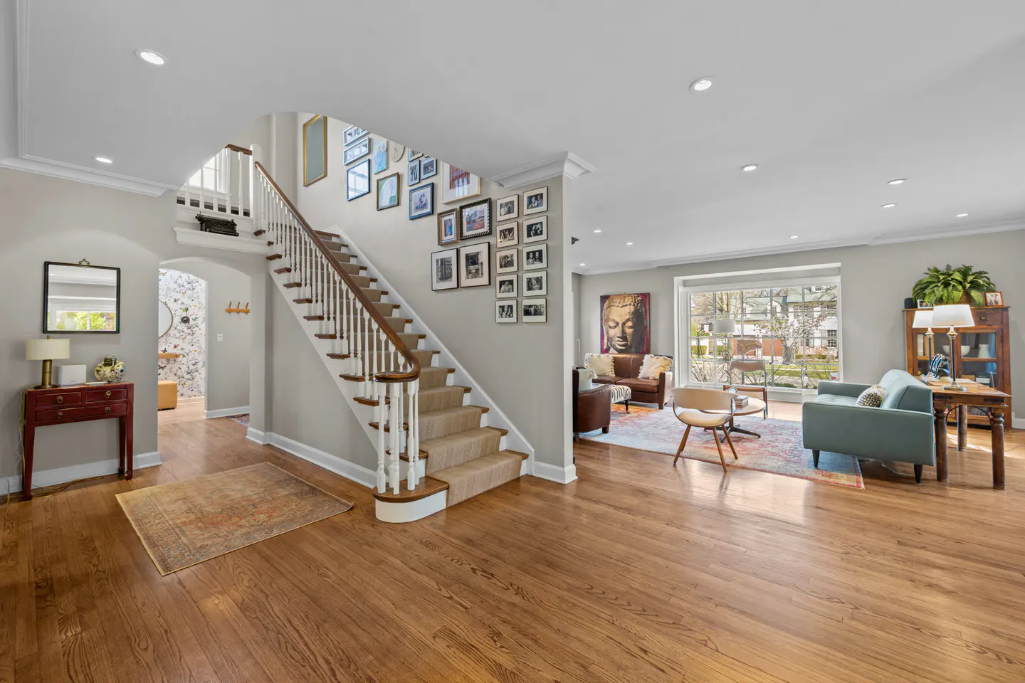 Bright foyer with hardwood floors, staircase with gallery wall, and living room with large window.