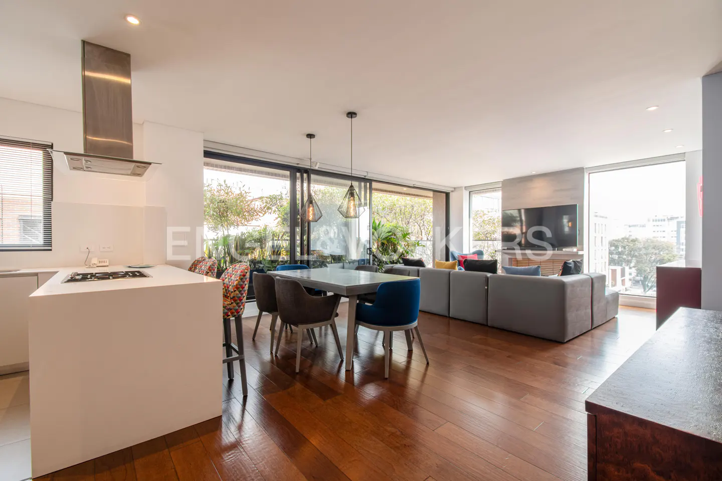 Open-concept living space with wood floors, a kitchen island, dining table, and gray sectional sofa. Large windows offer city views.