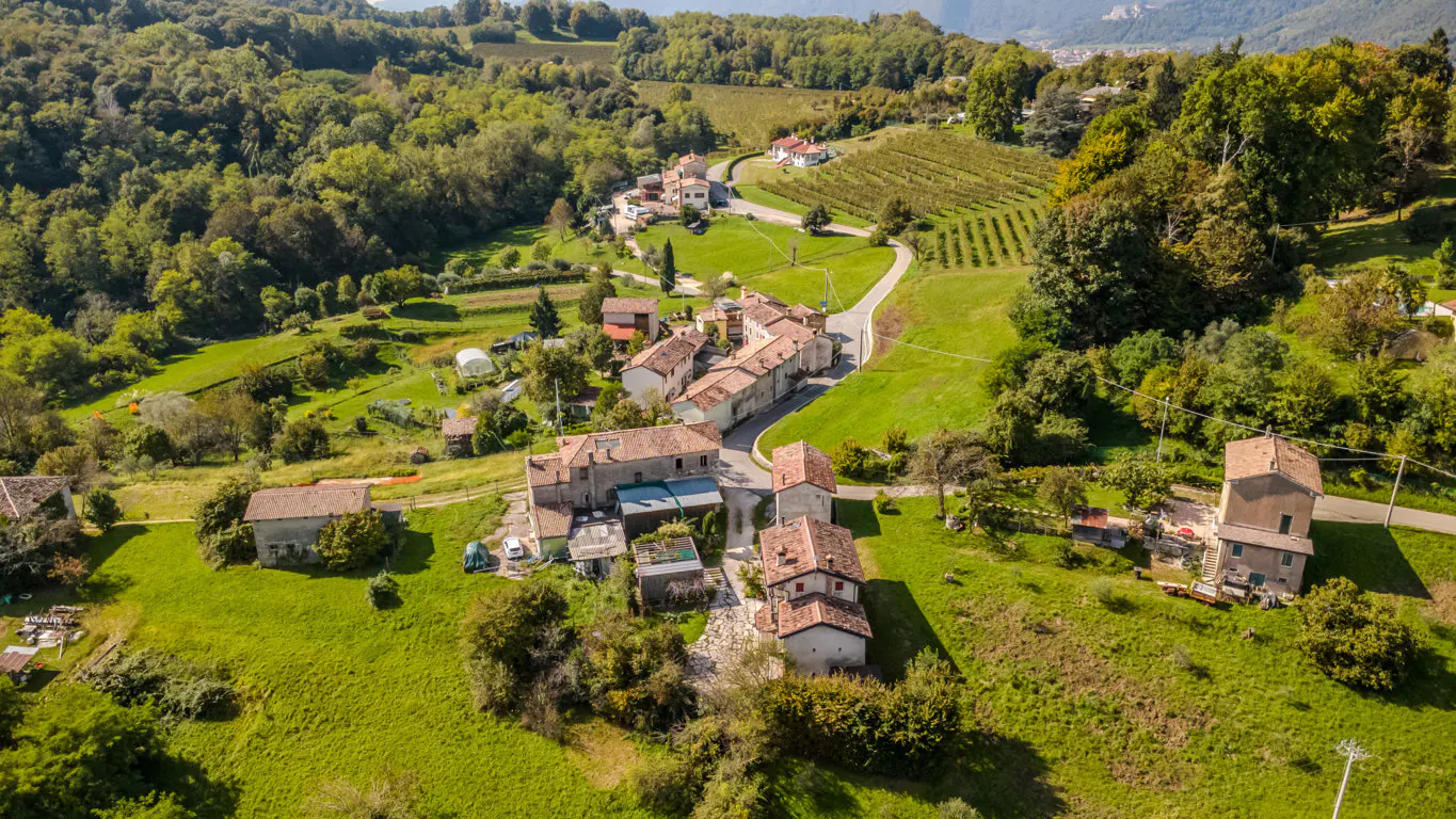 Aerial view of a rural Italian village with red-tiled roofs, surrounded by green hills, trees, and vineyards.