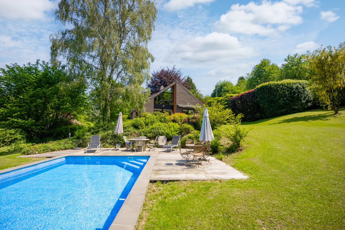 A blue tiled pool sits in a green lawn, with a house, trees, and patio furniture in the background.