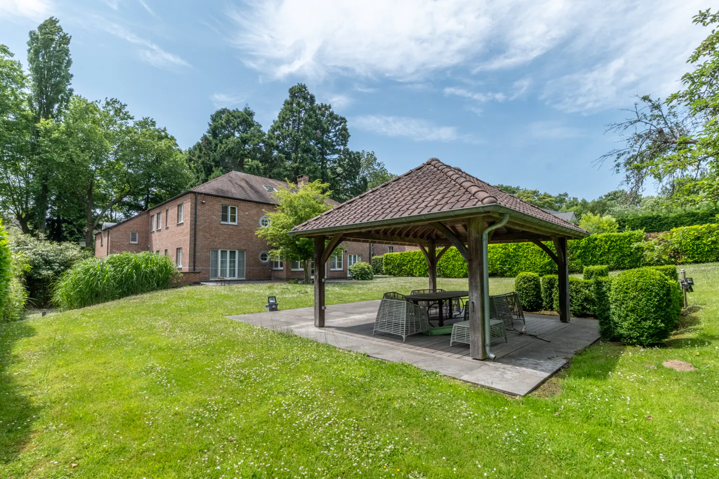 A backyard with a wooden gazebo, table, and chairs, surrounded by green grass, trees, and a brick building under a blue sky.