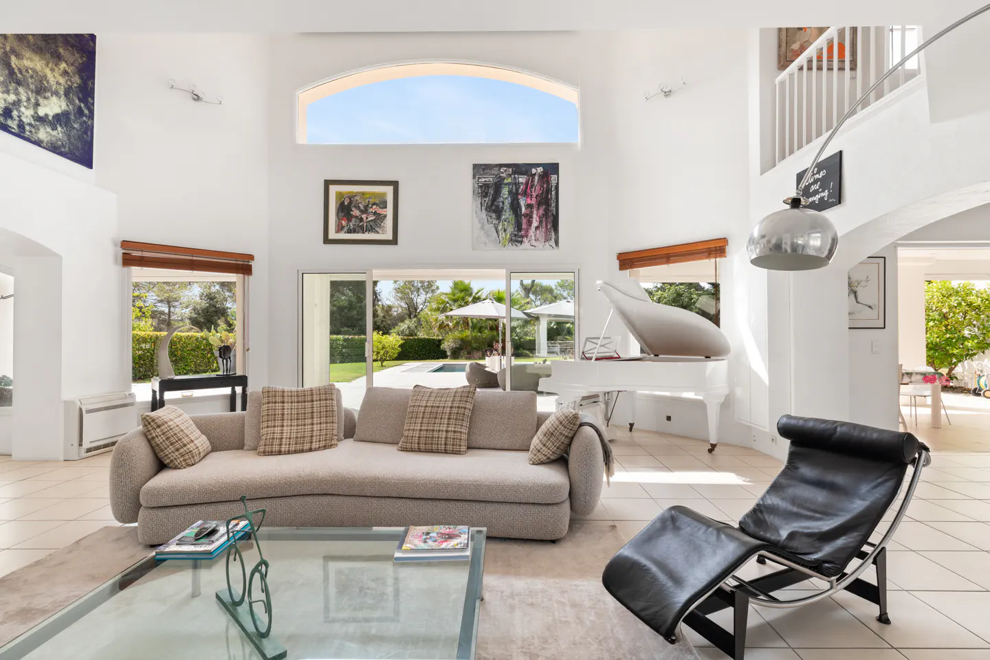 Bright living room with white walls, a beige sofa with plaid pillows, a white piano, and a black lounge chair. Outside is a pool and greenery.