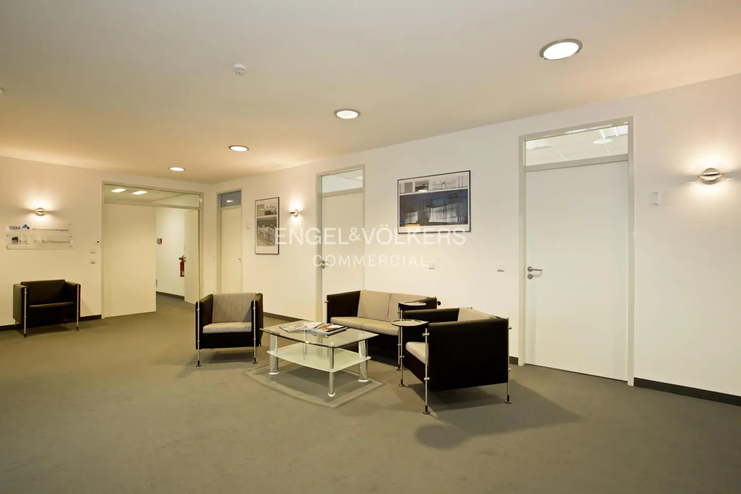A commercial lobby with a gray carpet, white walls, and modern furniture. A glass table sits in front of a sofa and chairs.