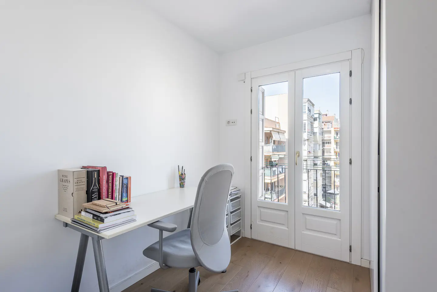 Bright, white home office with a desk, books, and a gray chair. Double doors open to a balcony with city views.