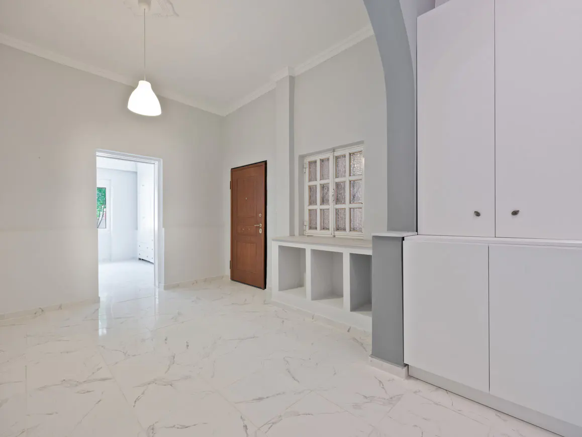 Bright, empty room with white marble floors, white walls, and a brown door. A white pendant light hangs from the ceiling. Built-in white cabinets are on the right.