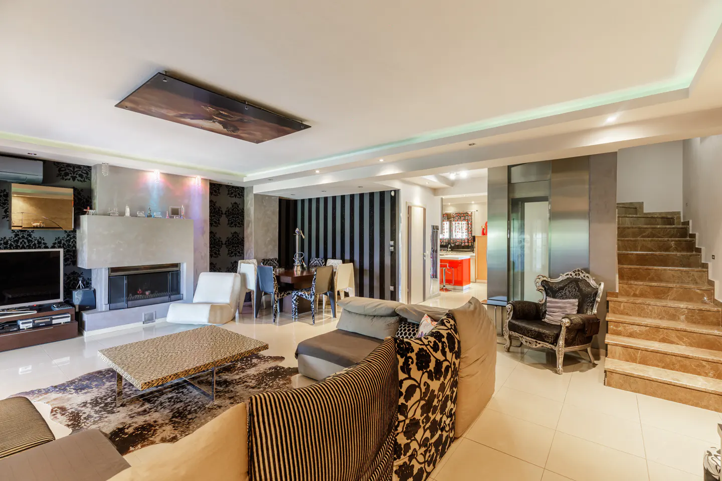 A modern living room with a fireplace, patterned wallpaper, and a dining area. A staircase is visible on the right.