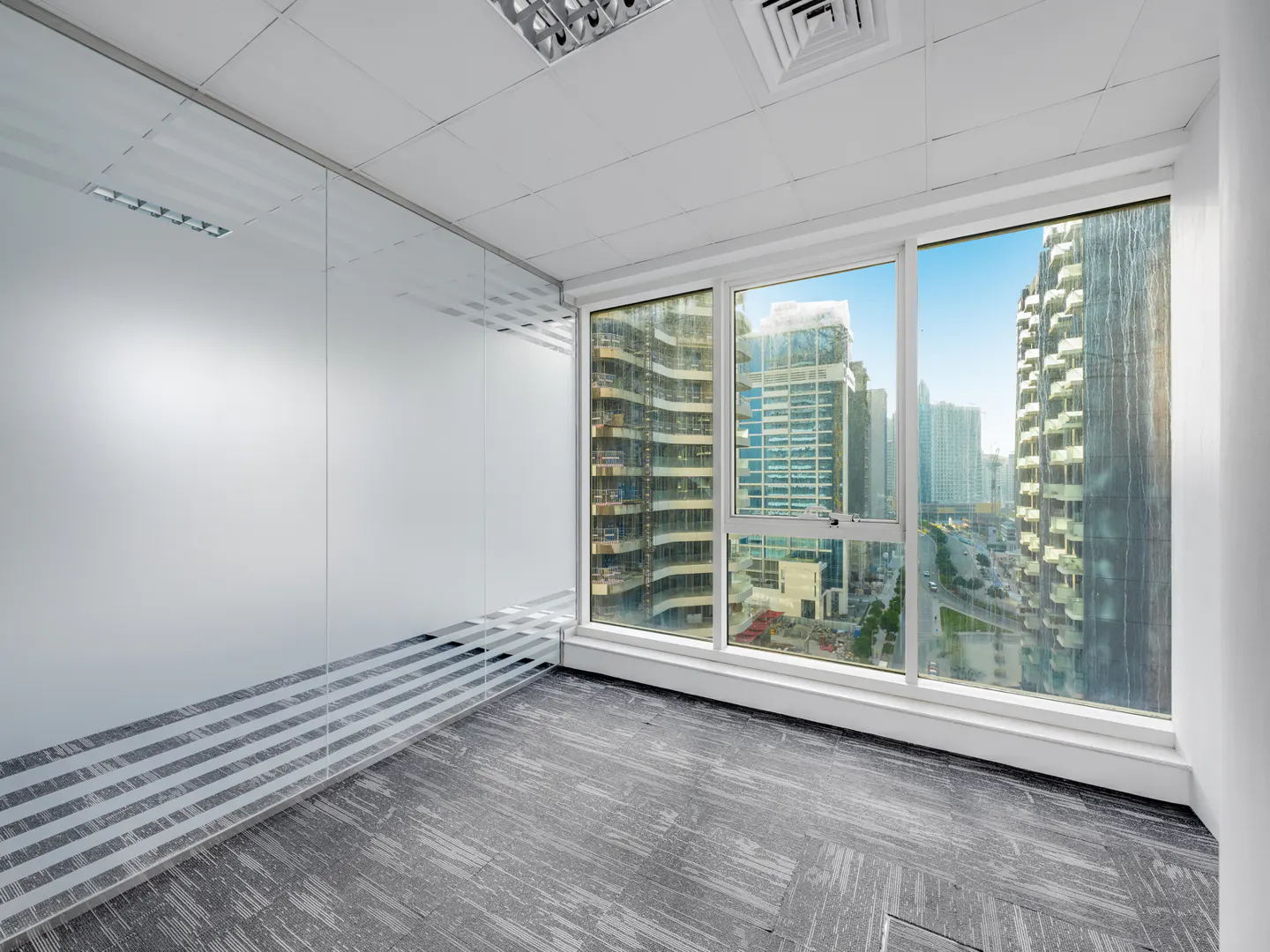 Empty office space with gray carpet, white walls, and a large window showing a city skyline with tall buildings.