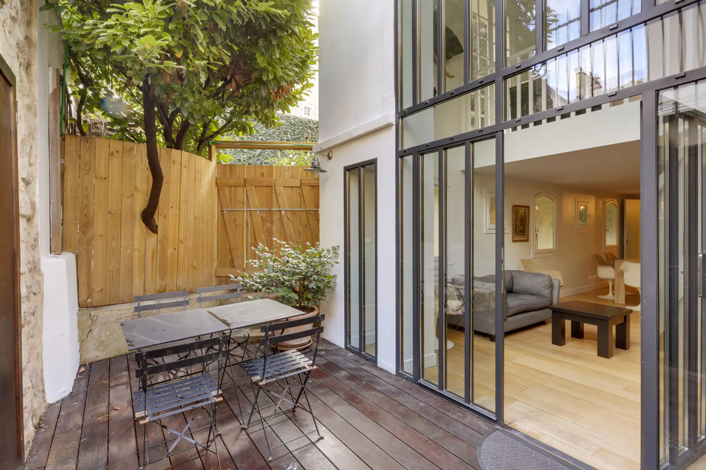 Outdoor patio with wood deck, table, and chairs. Large glass doors open to a living room with gray sofa. Wooden fence and tree in background.