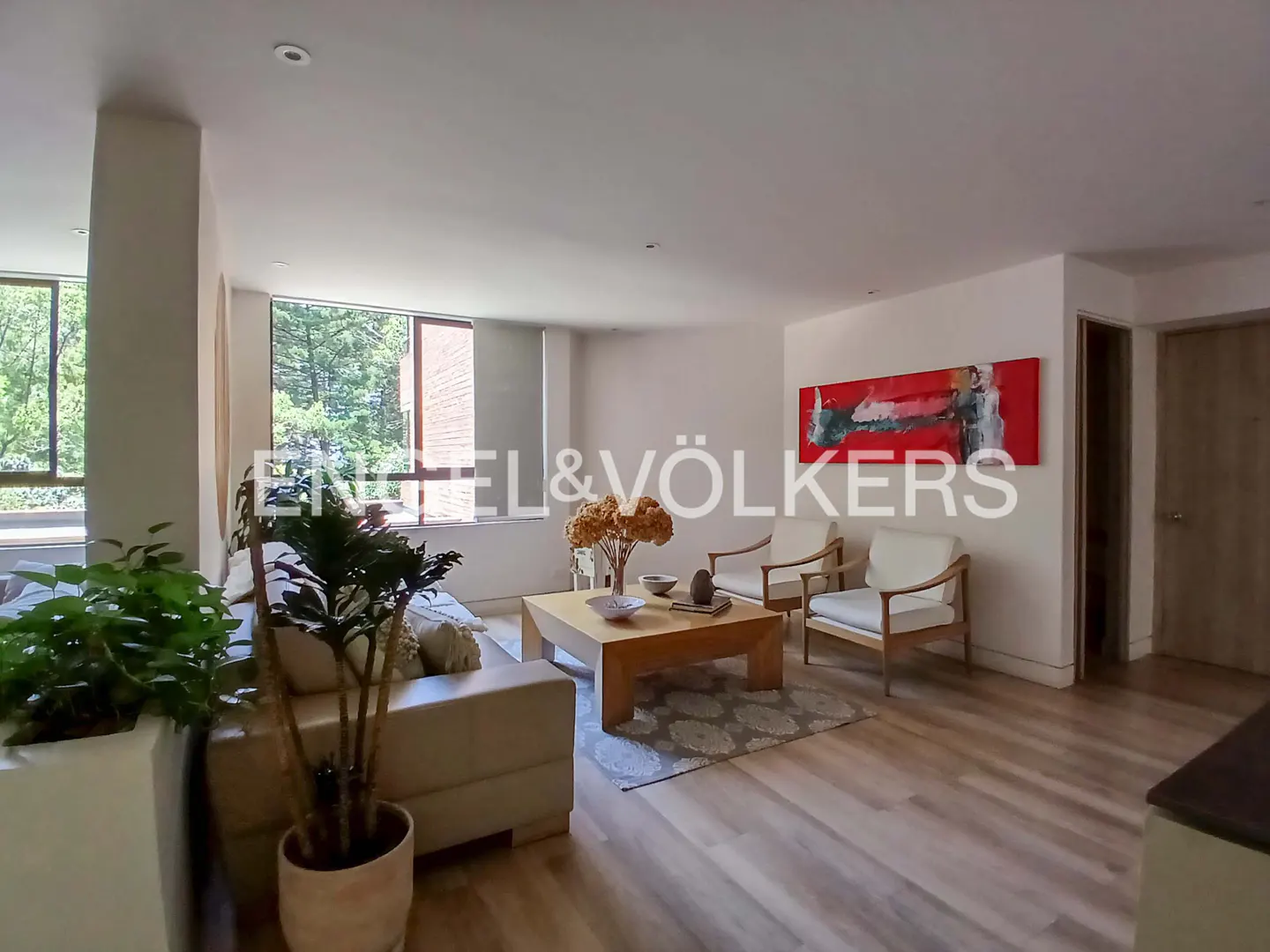 Bright living room with wood floors, beige sofa, two white chairs, and a wood coffee table. Large windows overlook trees. Red abstract art on the wall.