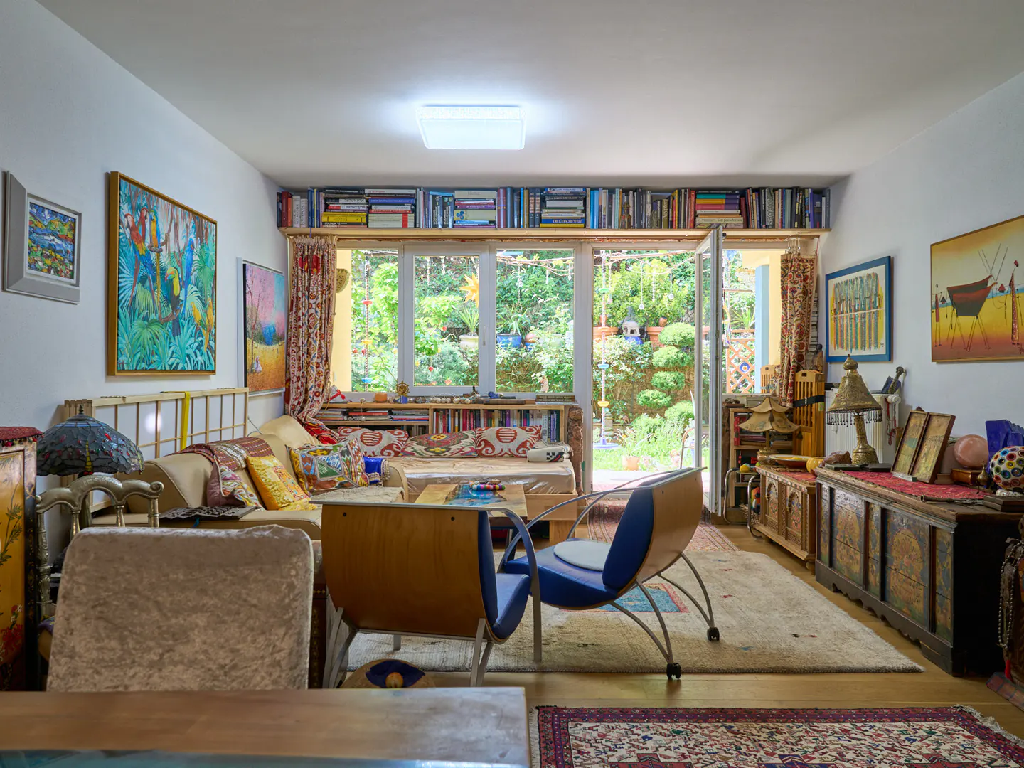 Bright living room with eclectic decor, including colorful art, books, and a view of a lush green garden through open doors.