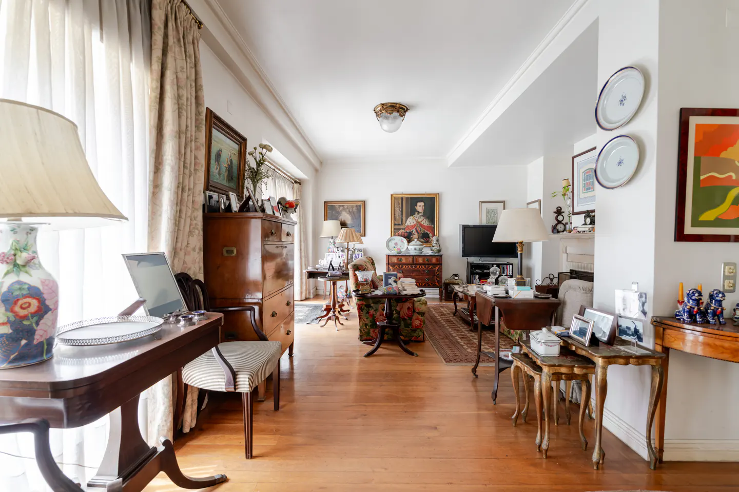 A bright living room with hardwood floors, white walls, and antique furniture. Lamps, paintings, and decorative plates add character.