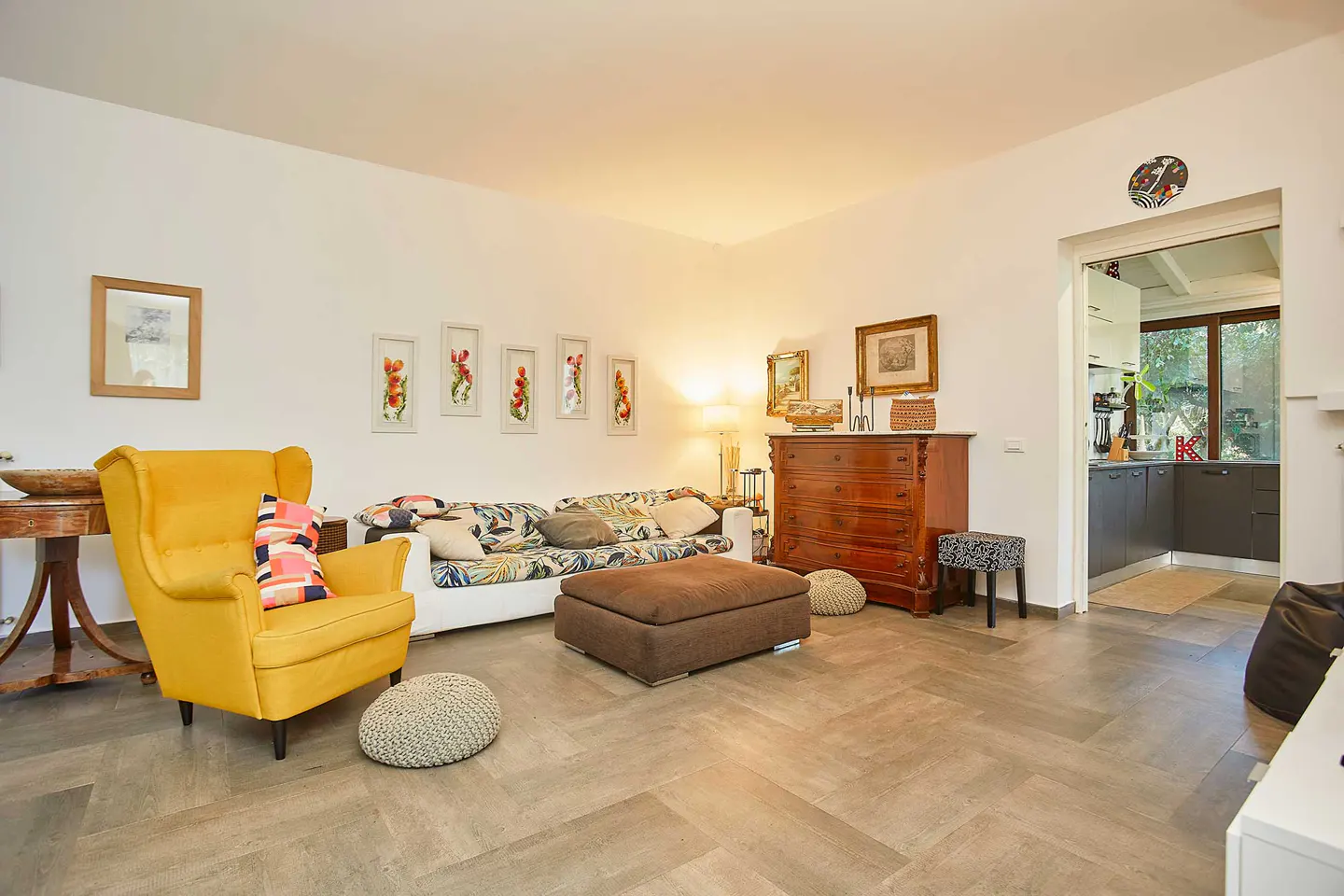 Bright living room with a yellow armchair, patterned sofa, brown ottoman, and wooden dresser, leading to a kitchen.