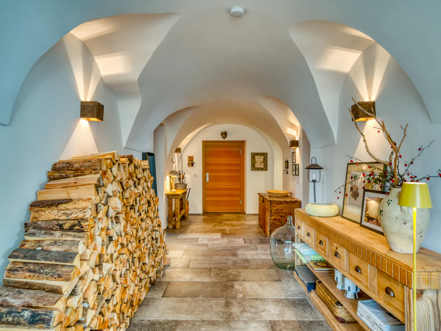Hallway with arched ceiling, stone floor, and wood accents. Firewood stacked on the left, wooden console table on the right.