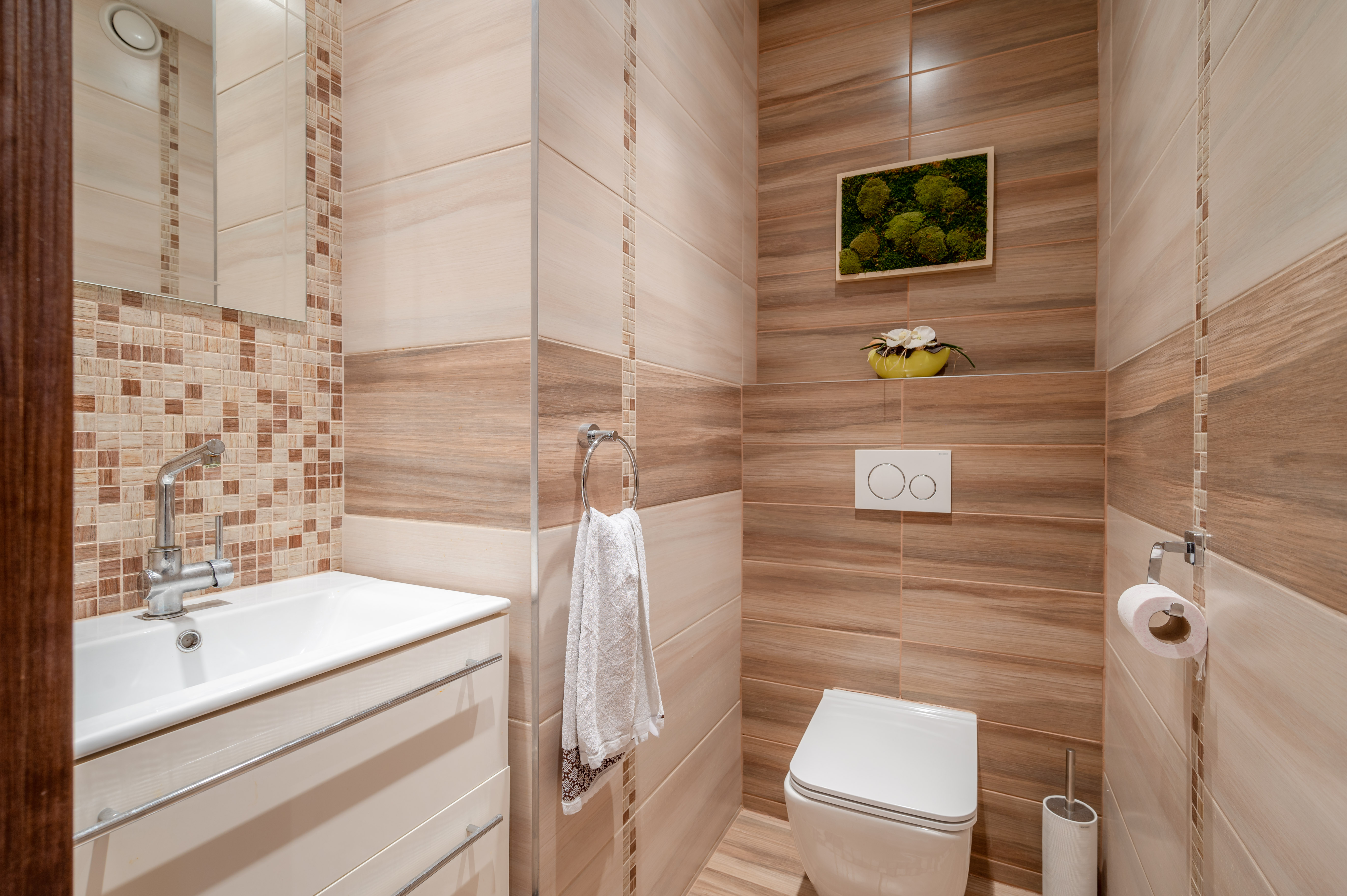 A modern bathroom with beige and brown tiles, a white toilet, sink, and a framed moss art piece on the wall.