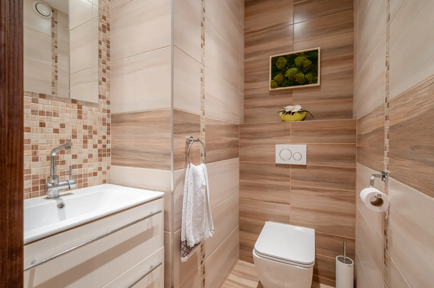 A modern bathroom with beige and brown tiles, a white toilet, sink, and a framed moss art piece on the wall.