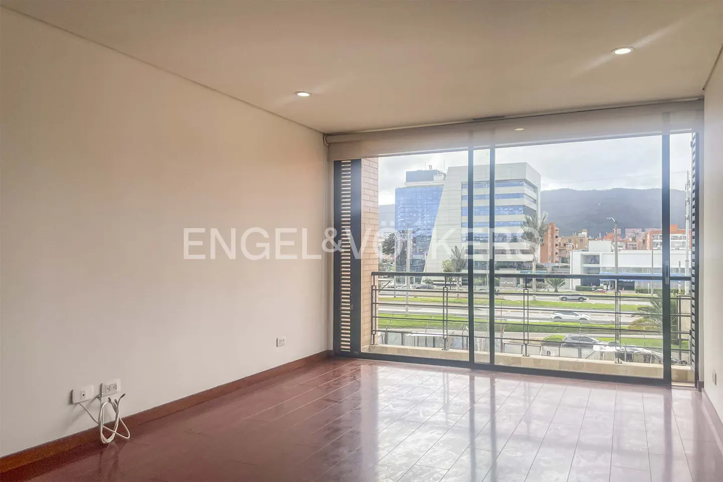 Empty room with hardwood floors and large windows overlooking a city street and buildings. Balcony with black metal railing. Cream colored walls.