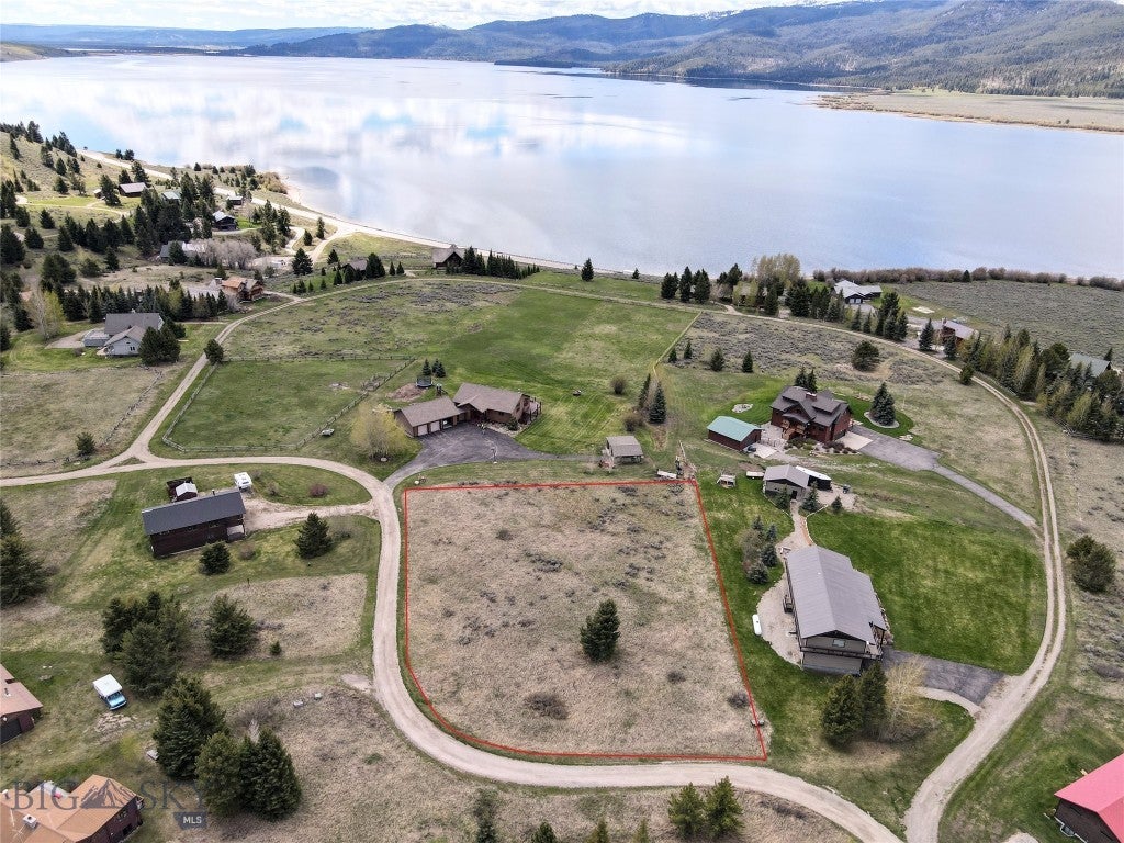 Aerial view of a vacant, grassy lot outlined in red, situated in a rural lakeside community with mountains in the distance. [AI-generated text]