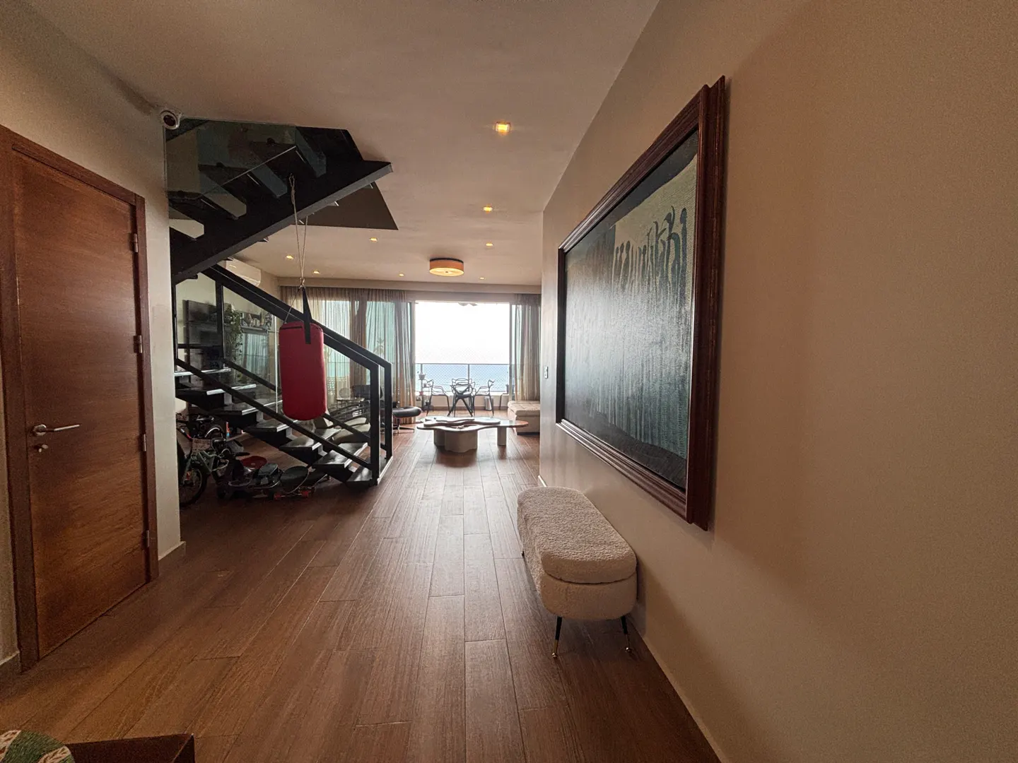 Hallway with wood floors, a staircase with a red punching bag, and a painting on the wall. Ocean view in the background.