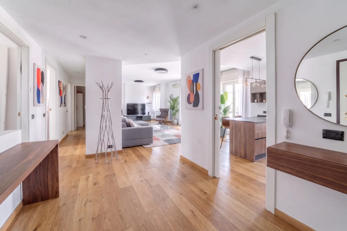 Bright apartment hallway with wood floors, white walls, and abstract art. Living room and kitchen visible in the background.