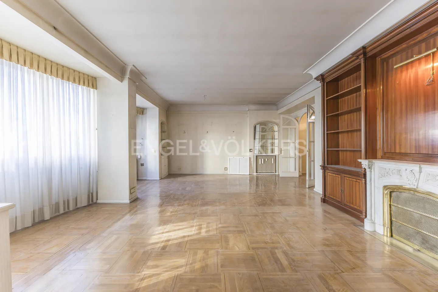 An empty room with parquet floors, white walls, and a large window with sheer curtains. A wooden bookcase and fireplace are on the right.