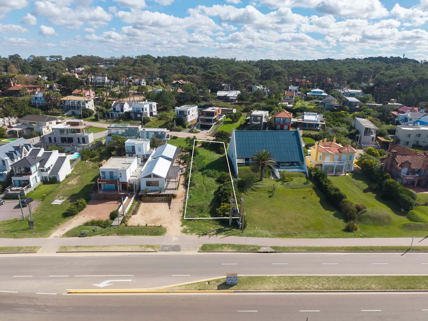 Aerial view of a vacant lot outlined in white, surrounded by houses and trees under a blue sky with white clouds. A road runs in front.