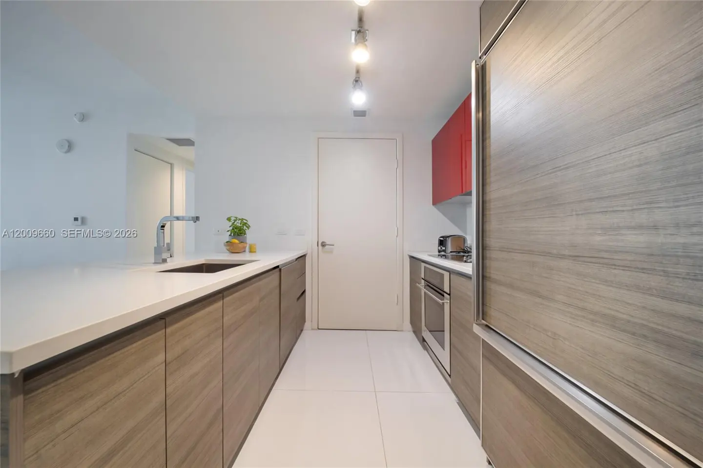 A modern kitchen with white walls, white tile floors, and wood-grain cabinets. A white countertop and stainless steel appliances are visible.
