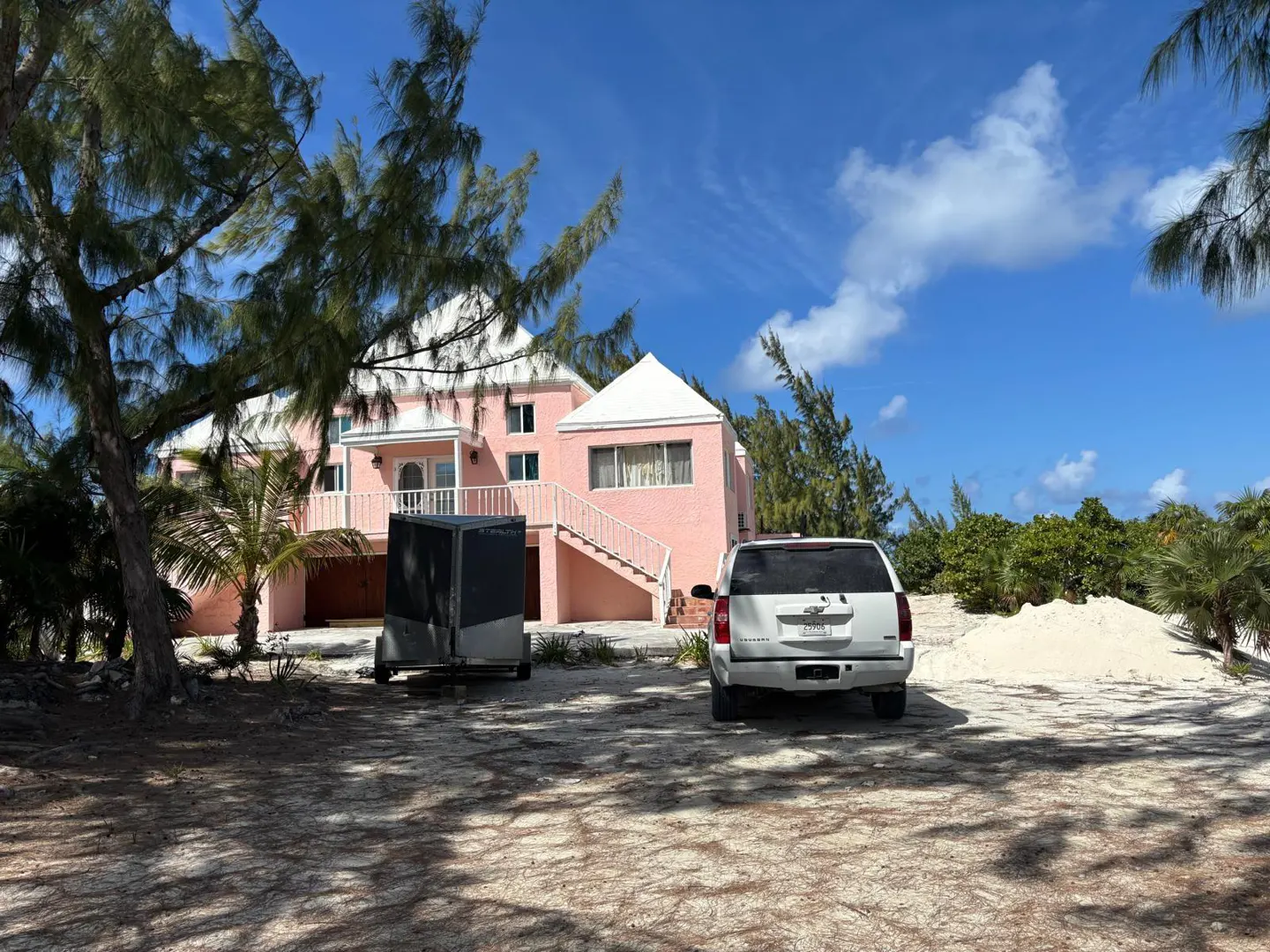 Pink house with white trim, stairs, and roof. A white SUV and trailer are parked in the sandy yard under a blue sky.