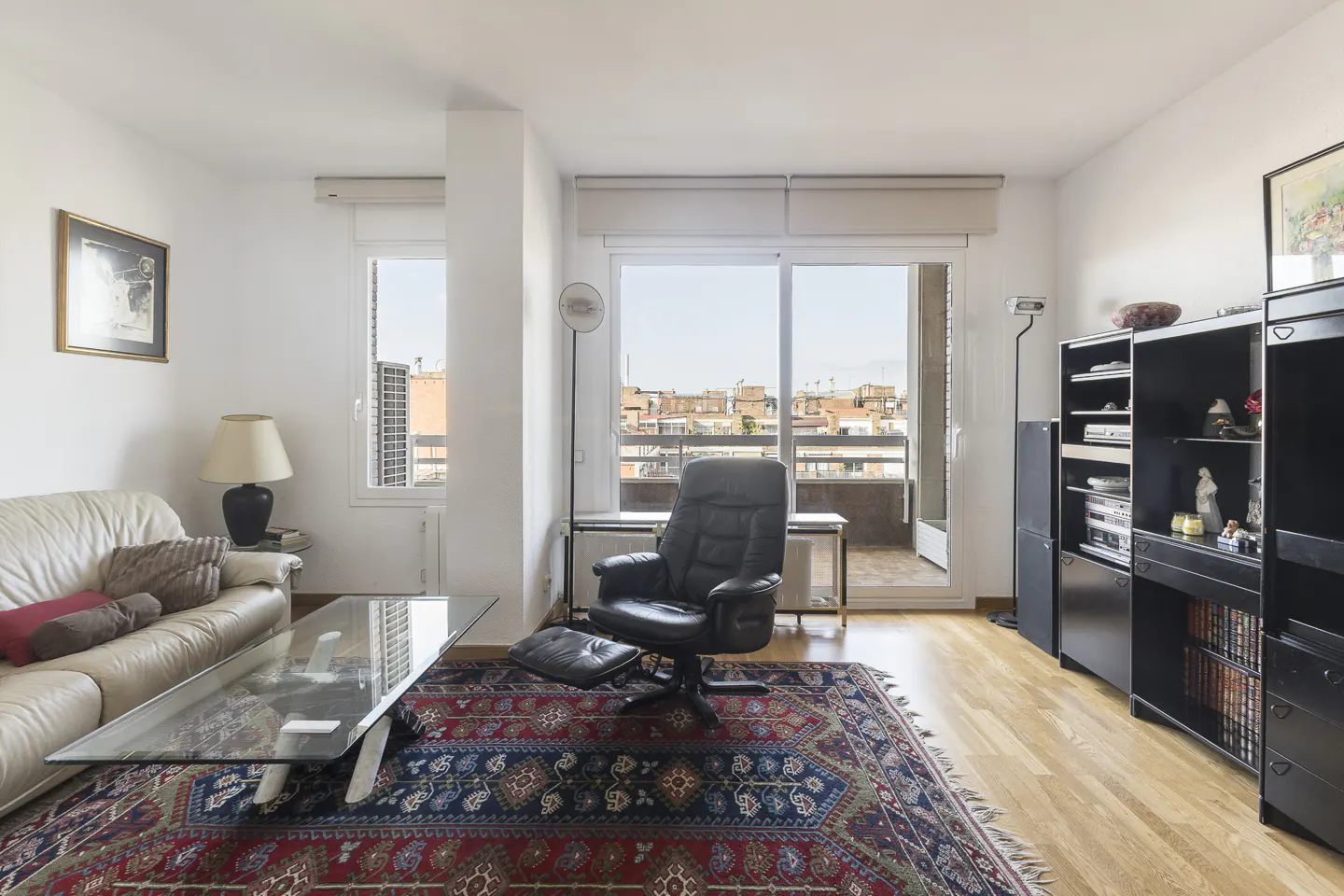 Bright living room with white walls, a leather sofa, a glass coffee table, and a black leather chair on a red and blue patterned rug. Balcony view.