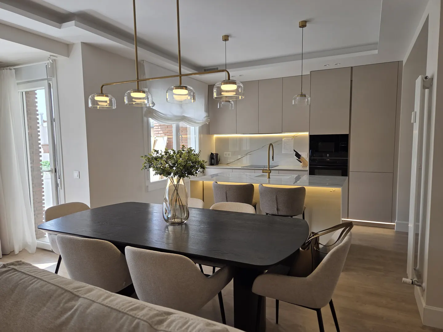 Modern dining area with a dark wood table, cream chairs, and a sleek kitchen with gold accents and warm under-cabinet lighting.