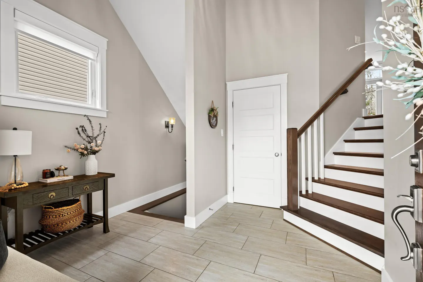 Entryway with beige walls, tile floor, and a dark wood console table with a lamp and flowers. Stairs with white risers and dark treads lead up. White door and window.
