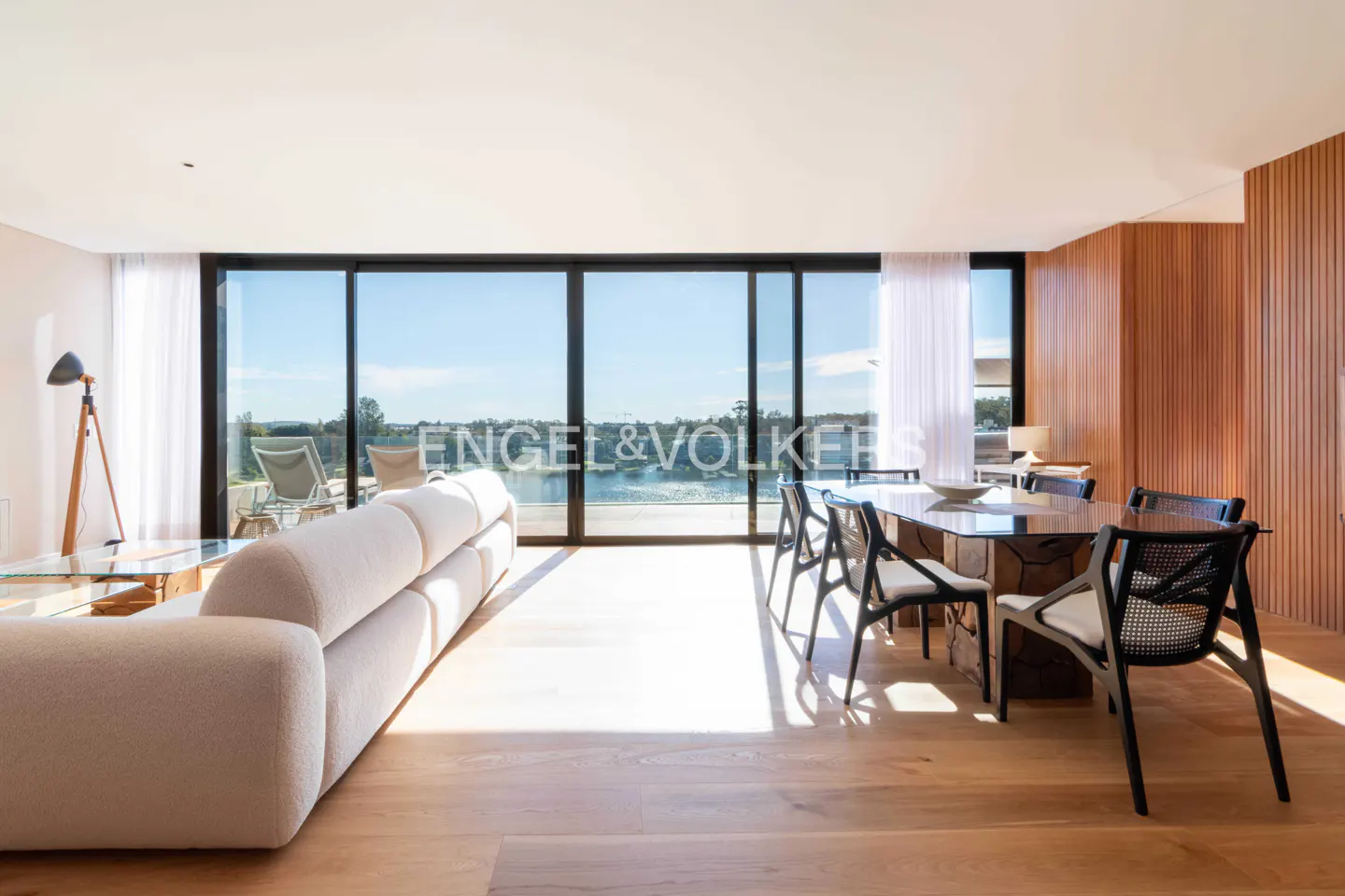 Modern living room with a cream sofa, dining area, and floor-to-ceiling windows overlooking a sunny waterfront view.