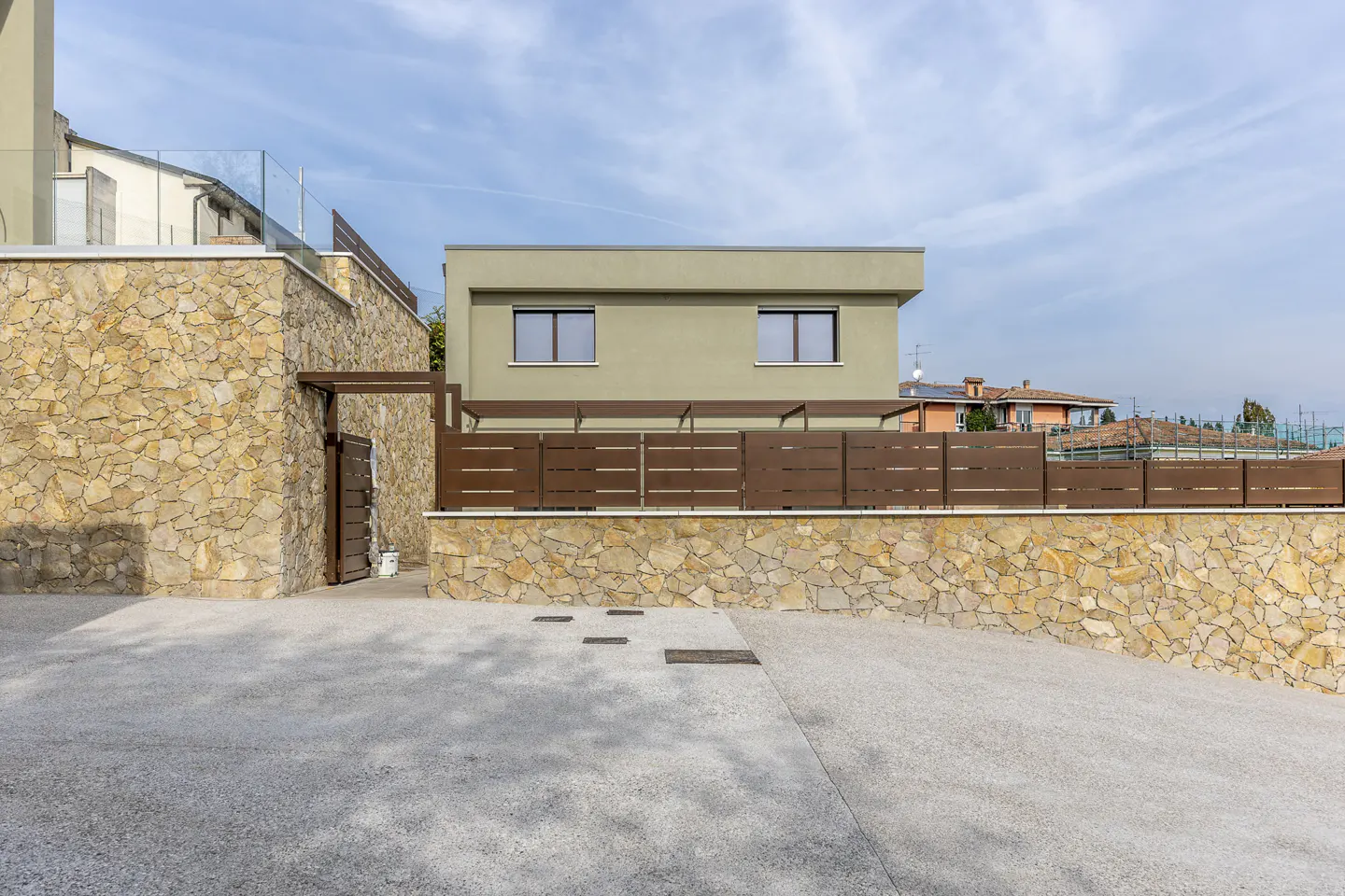 Exterior view of a two-story, light green house with brown trim and a stone wall. A concrete driveway leads to a wooden gate.
